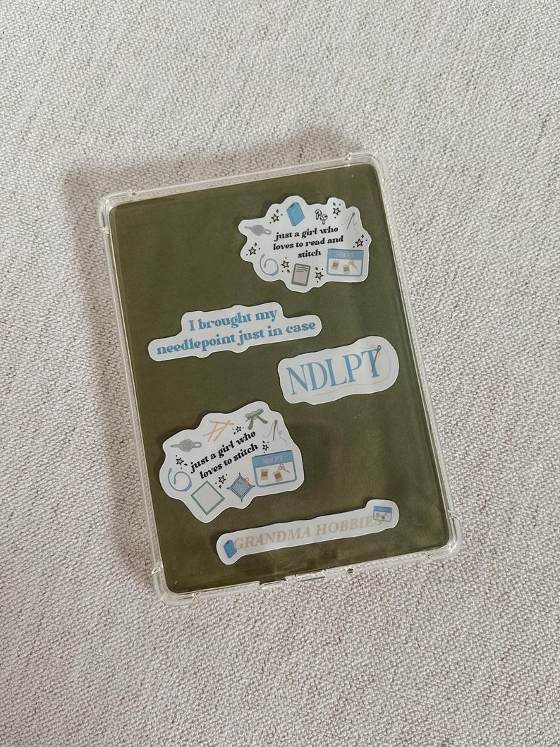May include: A clear protective case with a green interior, adorned with stickers. The stickers feature text like "just a girl who loves to read and stitch" and "I brought my needlepoint just in case."
