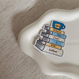 May include: A sticker featuring a stack of books with the words "Where You Lead I Will Follow" and a blue mug with the text "Luke's". The books have yellow and blue covers with white text. The sticker is on a white, wavy-edged tray.