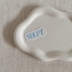 May include: A white, cloud-shaped ceramic tray with a glossy finish. A sticker with the text "NDLPT" in light blue is affixed to the tray. The tray is set against a neutral-toned, textured background.
