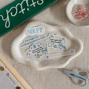May include: A collection of needlepoint supplies, including a green zippered pouch with "Stitch" embroidered in white, stickers, a small dish of pink thread, and light blue scissors. The stickers have text like "just a girl who loves to stitch".