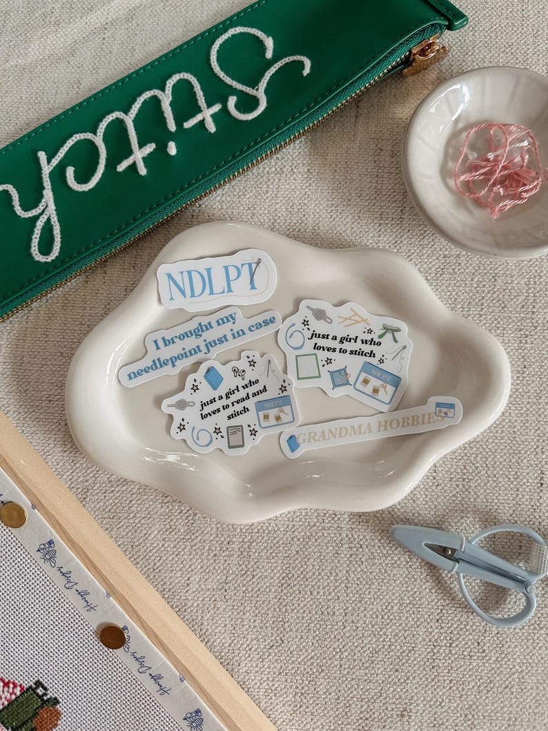May include: A collection of needlepoint supplies, including a green zippered pouch with "Stitch" embroidered in white, stickers, a small dish of pink thread, and light blue scissors. The stickers have text like "just a girl who loves to stitch".