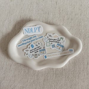 May include: A white, cloud-shaped ceramic dish holds several stickers with text and illustrations. The stickers feature phrases like "NDLPT," "I brought my needlepoint just in case," and "GRANDMA HOBBIES." Other stickers depict sewing-related items.