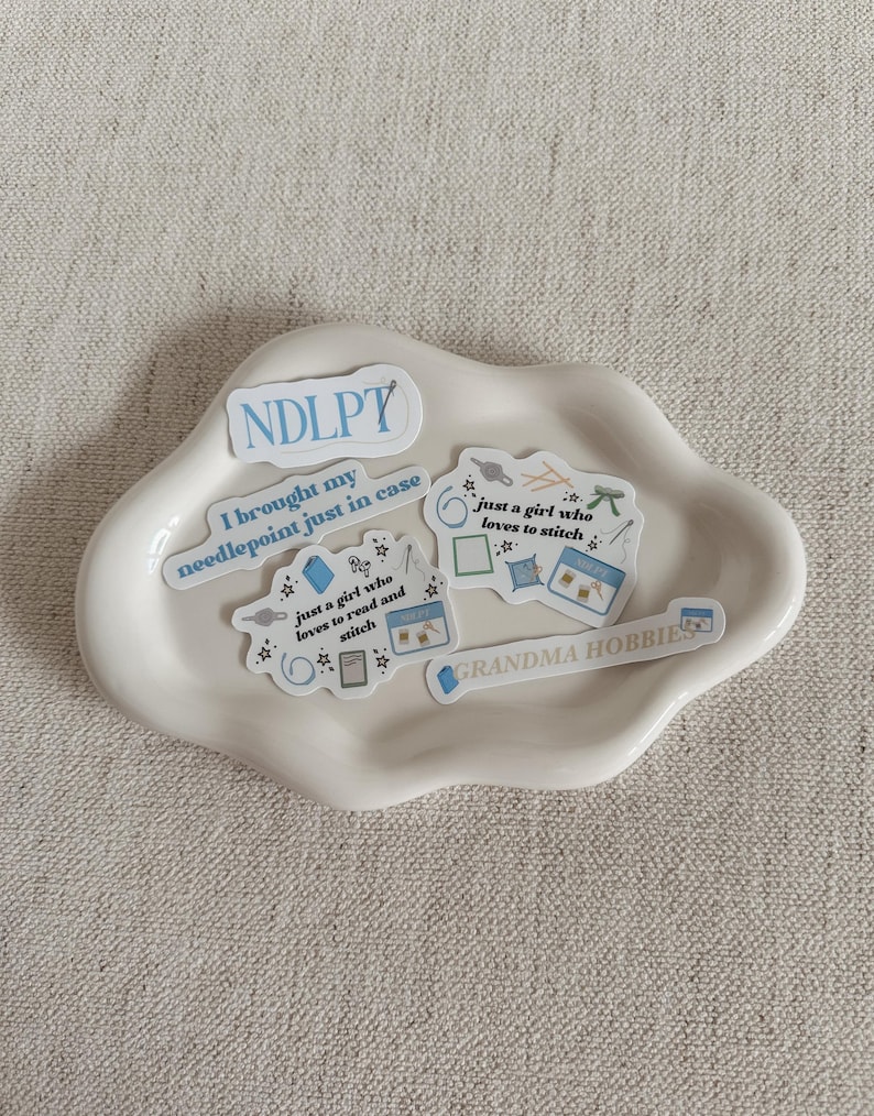 May include: A white, cloud-shaped ceramic dish holds several stickers with text and illustrations. The stickers feature phrases like "NDLPT," "I brought my needlepoint just in case," and "GRANDMA HOBBIES." Other stickers depict sewing-related items.