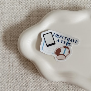 May include: A sticker with the text "I DON'T HAVE A TYPE" in blue, featuring a smartphone and headphones illustration. The sticker is on a white, wavy-edged ceramic dish, set against a neutral textured background.
