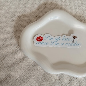 May include: A white, cloud-shaped ceramic dish holds a sticker with the text "I'm up late, cause I'm a reader." The sticker features a red lipstick kiss and a martini glass. The dish sits on a textured, neutral-toned surface.