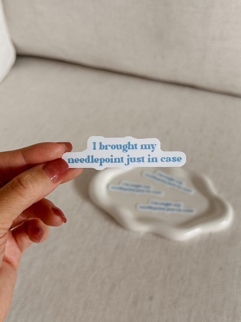 May include: A white sticker with the text "I brought my needlepoint just in case" in blue lettering. The sticker is held by a hand with painted nails, and there are several similar stickers on a white, wavy dish.
