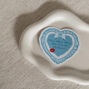 May include: A heart-shaped sticker with a light blue background and white lace trim. The sticker has the text "Please Please Please Take Me to the Bookstore" and a red lipstick kiss. The sticker is on a white, wavy-edged dish.