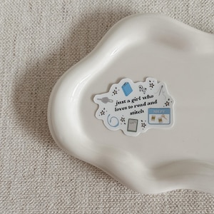 May include: A white sticker with the text "just a girl who loves to read and stitch" and illustrations of a book, scissors, and other craft supplies. The sticker is on a white, wavy-edged tray.