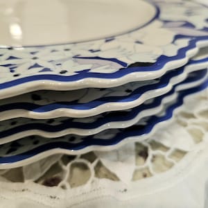 May include: A stack of white ceramic plates with a blue floral design and a wavy blue border. The plates are stacked on a white lace tablecloth, creating a classic and elegant aesthetic.