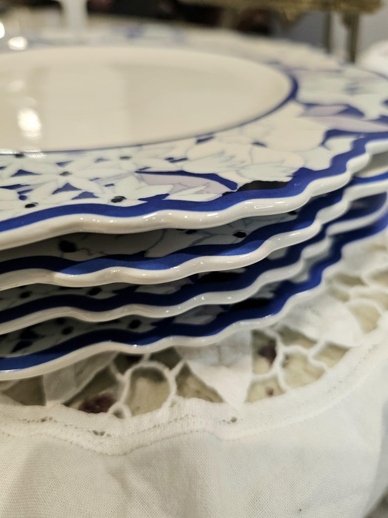 May include: A stack of white ceramic plates with a blue floral design and a wavy blue border. The plates are stacked on a white lace tablecloth, creating a classic and elegant aesthetic.
