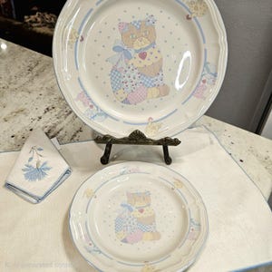 May include: A ceramic dinner plate and a smaller matching plate, both featuring a central patchwork-style cat illustration with a blue bow and heart pendant, surrounded by blue polka dots and a scalloped rim, accompanied by a folded linen napkin.