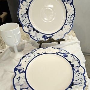 May include: Two white ceramic plates with a blue floral design border and a white wine glass with a face design. The plates are on a decorative stand. The plates have a scalloped edge.