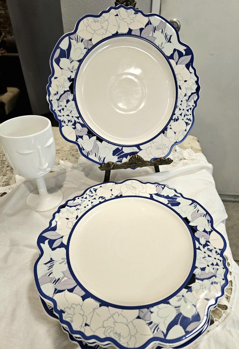 May include: Two white ceramic plates with a blue floral design border and a white wine glass with a face design. The plates are on a decorative stand. The plates have a scalloped edge.
