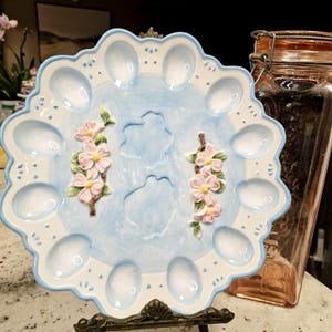May include: A round ceramic egg serving platter with a scalloped edge, featuring a light blue wash background, white border accents, and two raised, hand-painted floral branches with pink blossoms and green leaves centered on the plate.