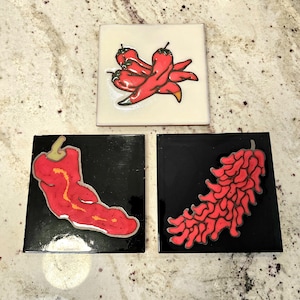 May include: Three square ceramic tiles featuring chilli pepper illustrations. One white tile displays a cluster of red chilli peppers. Two black tiles feature individual red chilli peppers, one with a smooth surface and one with a textured, bumpy surface.
