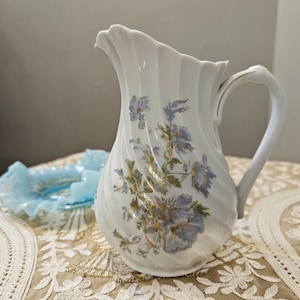 Puede incluir: Jarra de cerámica blanca con diseño en espiral y asa. La jarra está decorada con motivos florales azules y detalles dorados. Un plato azul claro con volantes se encuentra en el fondo.