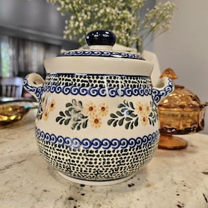 May include: A ceramic lidded jar with two side handles, featuring a cream-colored base decorated with blue spiral bands, green leaf patterns, and small yellow flowers, finished with a dark blue speckled texture on the bottom and lid knob.