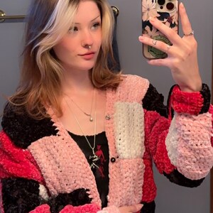 Könnte beinhalten: Eine Frau trägt eine mehrfarbige gehäkelte Strickjacke mit rosa, weißen und schwarzen Abschnitten. Die Strickjacke hat einen Knopfverschluss und ist aus weichem, flauschigem Garn gefertigt.