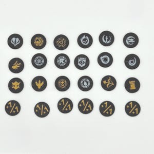 Peut inclure: Une collection de vingt-et-un jetons circulaires noirs avec divers symboles dorés et blancs. Les symboles incluent un cœur brisé, un crâne, un bouclier et un arc et des flèches. Les jetons sont disposés en grille sur une surface blanche.