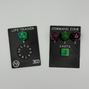 Puede incluir: Dos cartas de juego negras con texto y dados. Una carta dice "LIFE TRACKER" y tiene un dial y un dado verde. La otra carta dice "COMMAND ZONE" y tiene tres dados y la palabra "CASTS".