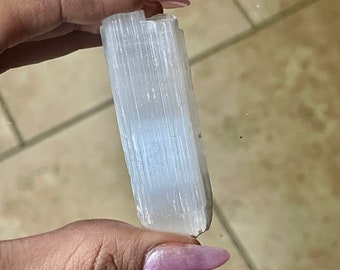 May include: A white selenite crystal stick with a striated texture. The crystal is held in a hand with pink nail polish.
