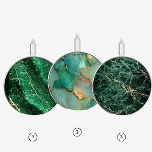Puede incluir: Tres bases de carga inalámbrica circulares, cada una con un patrón distinto inspirado en mármol verde con vetas doradas. Cada base tiene un conector de color plateado en la parte superior y está marcada con un círculo numerado: 1, 2 y 3.