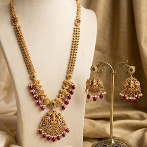 May include: Gold-toned necklace and earrings set with intricate detailing, red and green accents, and a pendant featuring a deity figure. The jewelry is displayed on a mannequin and stand, with a sign that reads "Temple Traditions - Heritage Collection."