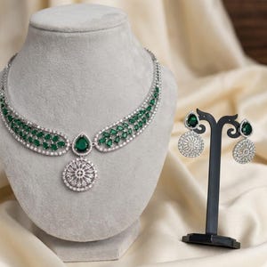 May include: A silver-toned necklace and earring set. The necklace features a central emerald-green teardrop-shaped stone and a circular pendant. The earrings have a similar design, with emerald-green stones and circular, sparkling accents. The jewelry is displayed on a gray mannequin head and a black stand.