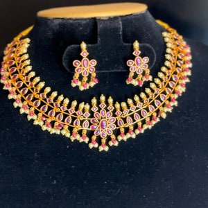 May include: A gold necklace with pink gemstones and pearl accents. The necklace has a delicate design with a floral motif. Matching earrings with pink gemstones and pearls are included.