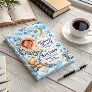May include: A hardcover baby memory book featuring a blue sky background with stars, a crescent moon, and a teddy bear. The cover displays a photo of a sleeping infant, the text Welcome to the world, Oliver James, September 18, 2026, 8:42 AM, 7 lb 6 oz, 20 inches.