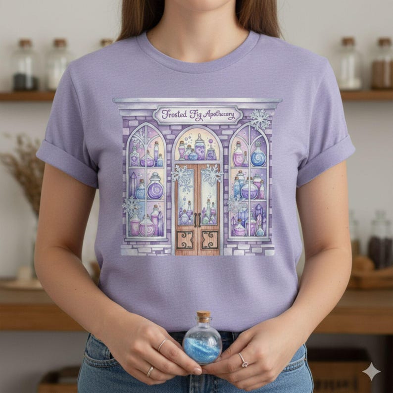 K&ouml;nnte beinhalten: Helllila T-Shirt mit einer Aquarellillustration eines Gesch&auml;fts namens "Frosted Fig Apothecary". Das Gesch&auml;ft hat gew&ouml;lbte Fenster mit Zaubertr&auml;nken in Blau, Lila und Rosa. Eine kleine Glasflasche mit blauem Glitzer wird in den H&auml;nden gehalten.