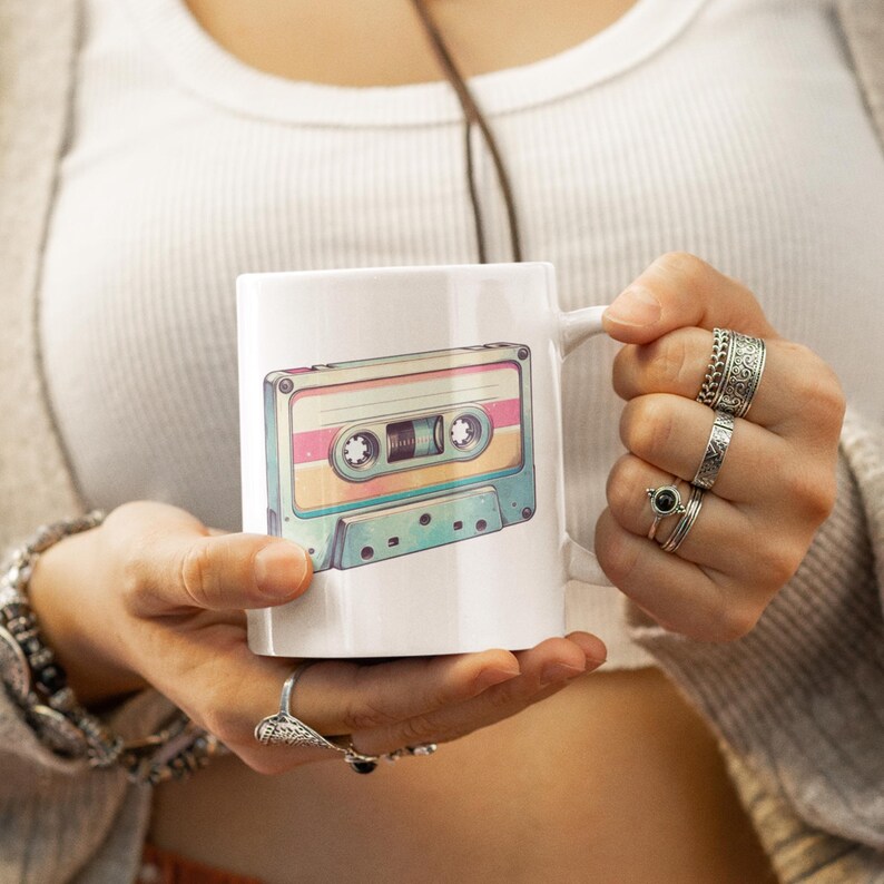 May include: White ceramic mug with a vintage cassette tape design. The cassette tape is illustrated in a retro style with pink, blue, and yellow colors.