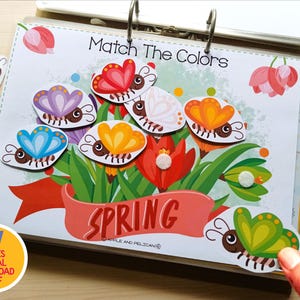 May include: A colorful educational activity book with the text "Match The Colors" and the word "Spring". The image features butterfly cutouts in various colors, along with markers. The book is held open with metal rings.