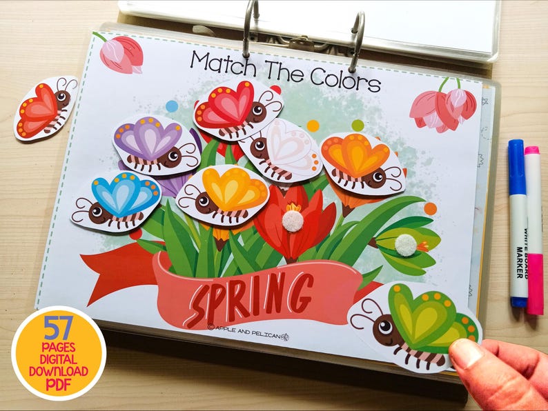May include: A colorful educational activity book with the text "Match The Colors" and the word "Spring". The image features butterfly cutouts in various colors, along with markers. The book is held open with metal rings.