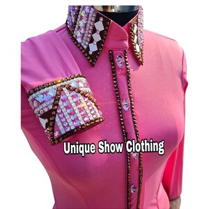 Może przedstawiać: Różowa koszula z długim rękawem z brązowymi i białymi zdobieniami z kryształków na kołnierzyku i mankietach. Koszula zapinana jest na guziki z przodu, a na przodzie nadrukowany jest napis "Unique Show Clothing".