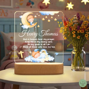 May include: A personalized night light with the name "Henry Thomas" and a prayer. The acrylic panel features illustrations of a baby on a crescent moon, clouds, stars, and a cross. The base is made of wood.