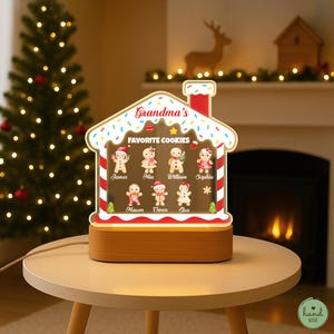May include: A gingerbread house-shaped night light with the text "Grandma's Favorite Cookies." The light features gingerbread people with names below them. The base is a warm brown color, and the house has red and white stripes.
