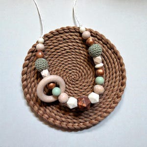 May include: A necklace with a variety of beads in neutral tones, including wood, white geometric shapes, and green crocheted spheres. The necklace features a wooden ring and is displayed on a woven, circular mat.