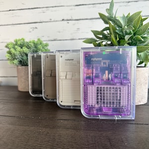 May include: Four clear plastic handheld game consoles. The consoles are in different colors: black, white, gray, and purple. The purple console has a visible circuit board.