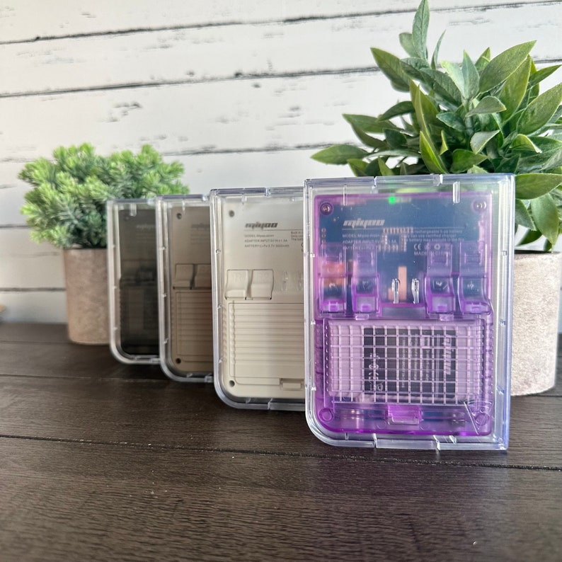 May include: Four clear plastic handheld game consoles. The consoles are in different colors: black, white, gray, and purple. The purple console has a visible circuit board.