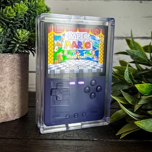 Könnte beinhalten: Eine blaue und schwarze Handheld-Videospielkonsole ist in einem durchsichtigen Plastikgehäuse eingeschlossen. Die Konsole hat einen Bildschirm, auf dem das Spiel "Paper Mario" mit dem Text "Press Start" und "2001 Nintendo Intelligent Systems" angezeigt wird.