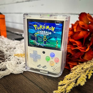 Könnte beinhalten: Handheld-Spielkonsole mit transparentem Schutzgehäuse. Der Bildschirm zeigt "Pokémon Emerald Version" mit grünem und blauem Hintergrund. Das Gerät hat ein cremefarbenes Gehäuse mit farbigen Knöpfen und einem Steuerkreuz.