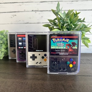 May include: Four clear plastic cases with different colored Game Boy Advance handheld game consoles inside. The consoles are black, white, purple, and brown. The Pokemon FireRed Version game is displayed on the black console.