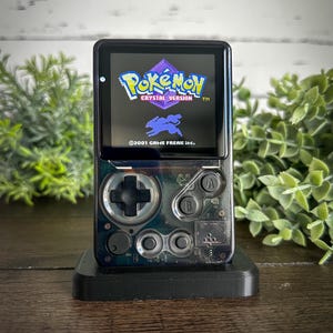 May include: A black handheld gaming device displaying the "Pokémon Crystal Version" game. The device has a clear, transparent design with visible internal components. It rests on a black stand, with a wooden surface in the background.