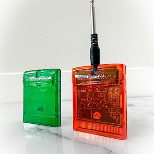 May include: Two Game Boy Color cartridges, one green and one orange, are displayed. The orange cartridge has a black cable and antenna plugged in. The cartridges are on a white surface.