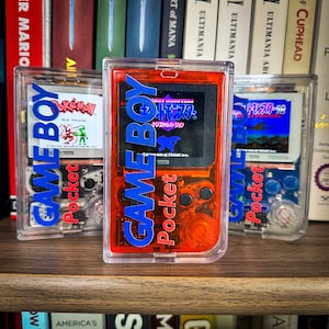 Könnte beinhalten: Drei transparente Plastikhüllen mit Game Boy Pocket Konsolen darin. Die Konsolen sind rot, blau und klar. Die Bildschirme zeigen verschiedene Spiele an, darunter Pokemon Red Version, Grey Monsters und Grey Monsters Crystal Version. Der Text auf den Konsolen lautet "GAME BOY Pocket".