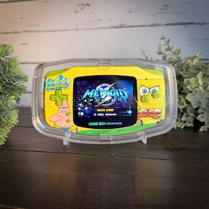 Könnte beinhalten: Ein gelbes Game Boy Advance Handheld-Konsolen mit einer transparenten Schutzhülle. Der Bildschirm zeigt das Spiel "Metroid Fusion" mit dem Text "Press Start" und "Game Boy Advance". Die Konsole ist mit SpongeBob Schwammkopf-Figuren dekoriert.