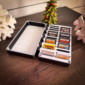 May include: A black and white hinged case, open to reveal a white interior and a section for game cartridges. The case holds several game cartridges, including "Brain Age" and "Zelda". A small Christmas tree is in the background.