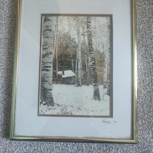 May include: Framed print of a winter forest scene with birch trees and a small cabin. The artwork is set in a gold-colored frame and features a snowy landscape with a touch of autumn colors. Signed in the bottom right corner.