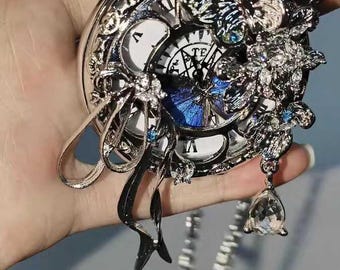 May include: Ornate silver pocket watch with a detailed floral and butterfly design. The watch face features Roman numerals and a blue butterfly accent. A delicate chain and crystal pendant complete the elegant aesthetic.